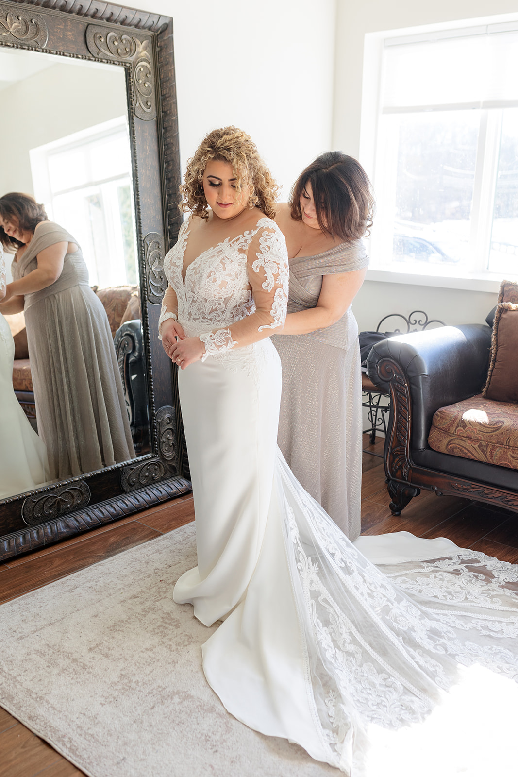 A bride is helped by mom to close up the back of her lace wedding dress in a mirror