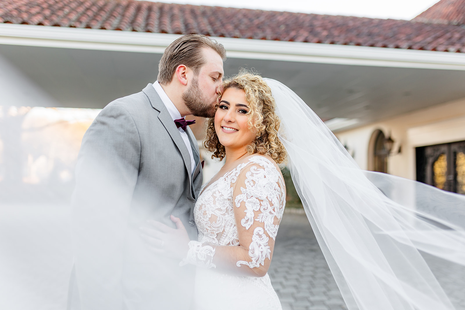 A groom in a grey suit kisses the head of his bride in a lace embroidered dress as the veil flows around them