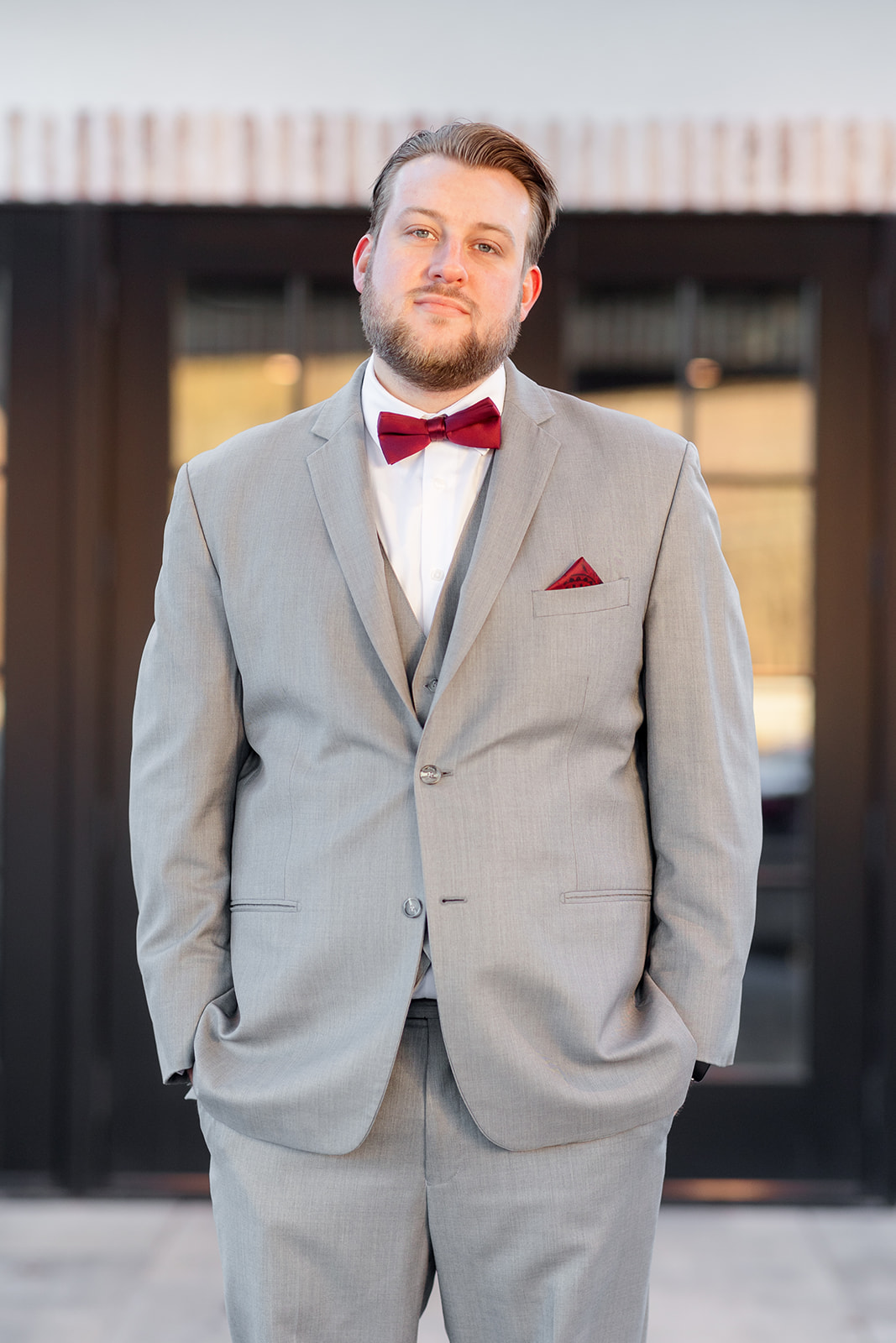 A groom stands with hands in the pockets of his grey suit with a red bowtie