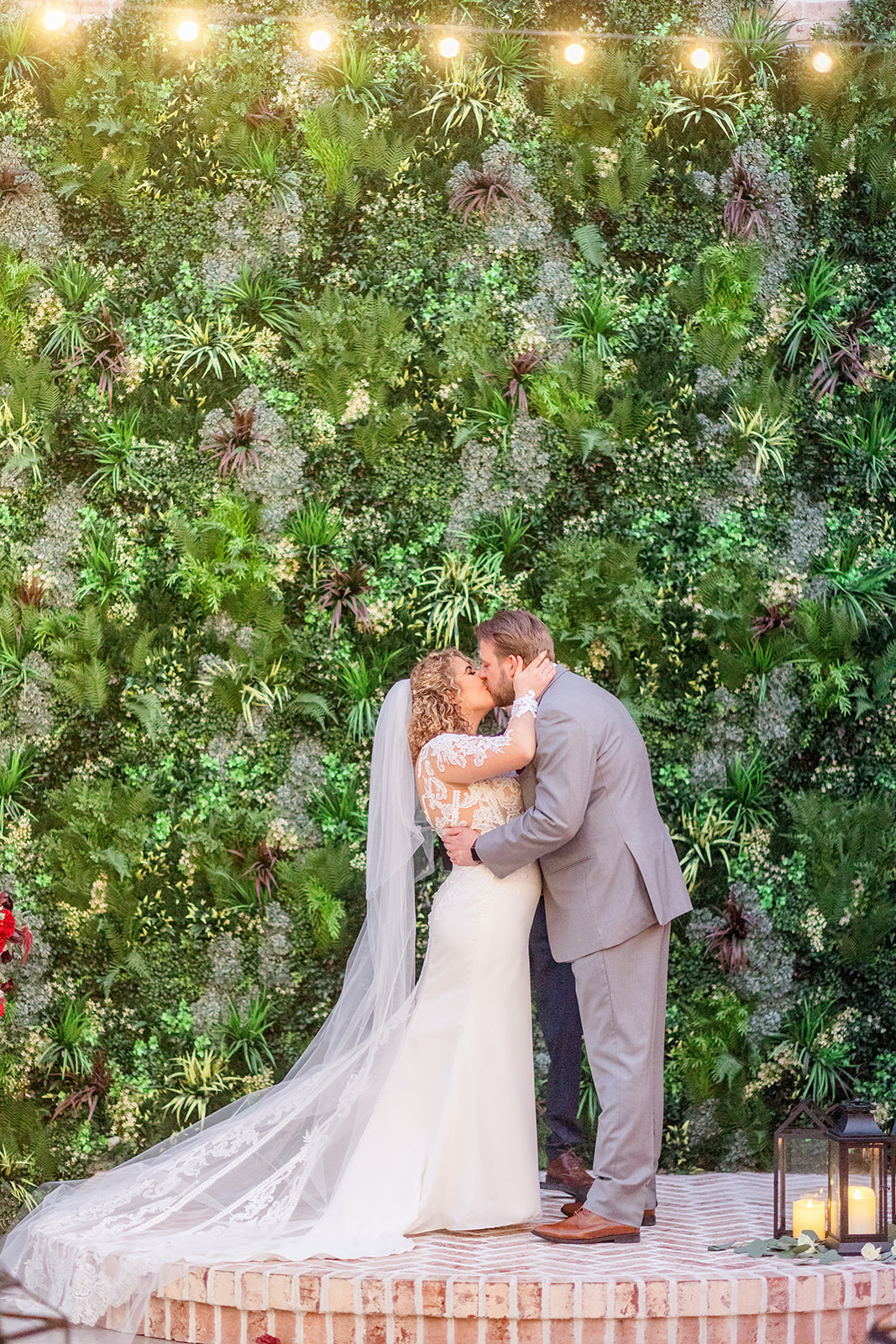Newlyweds kiss while standing in front of a greenery wall under market lights on a brick pedestal