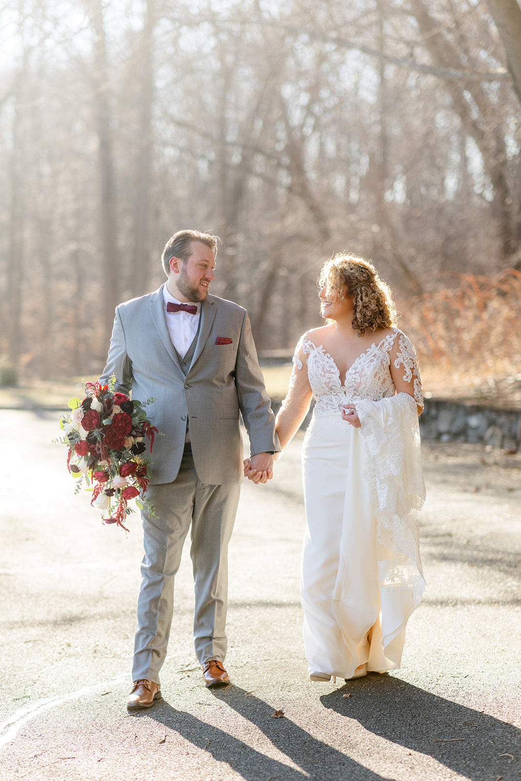 Newlyweds hold hands while the groom carries the large red bouquet and walking up a large sidewalk at sunset