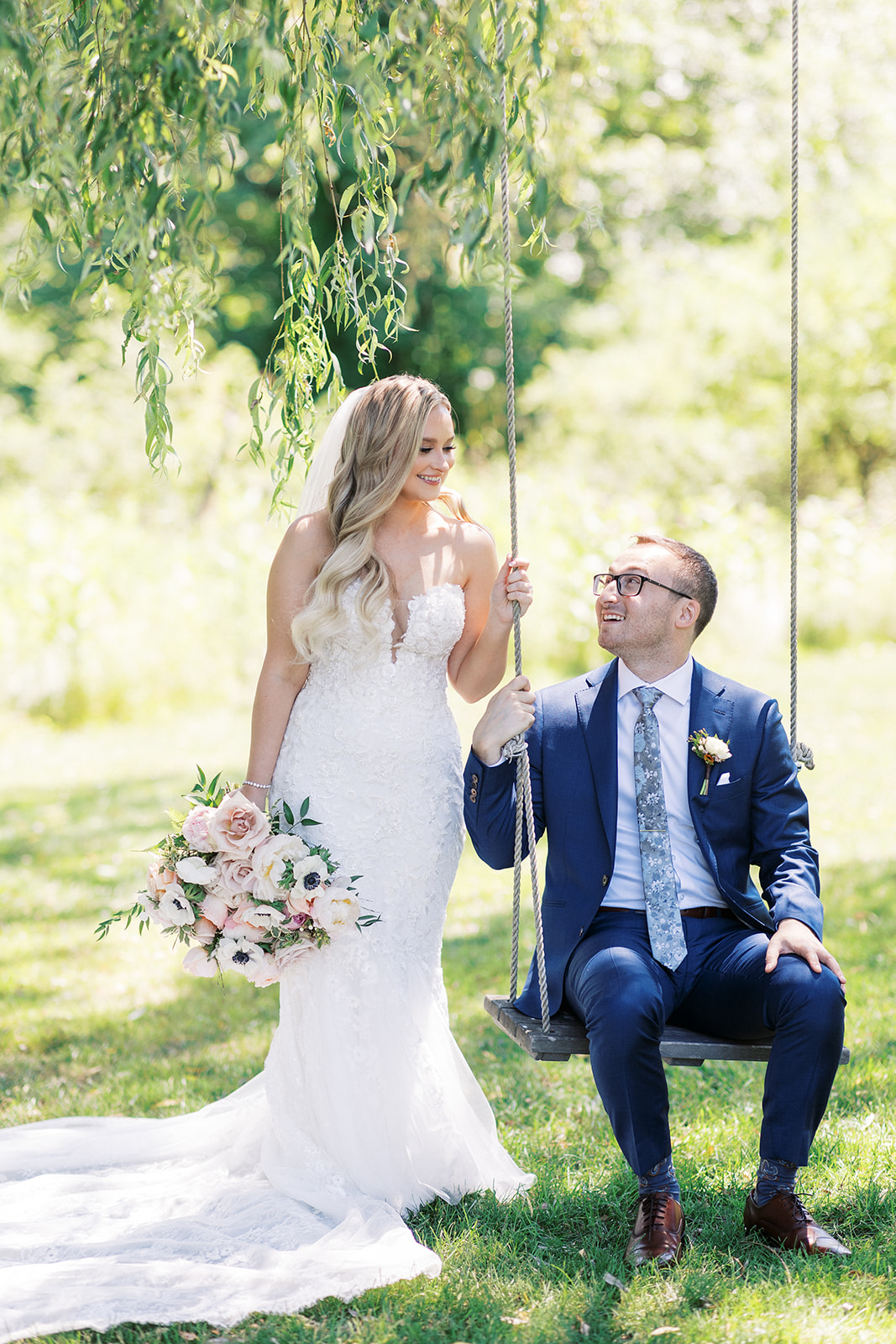 A groom in a blue suit sits on a tree swing as his bride leans on the rope smiling down at him