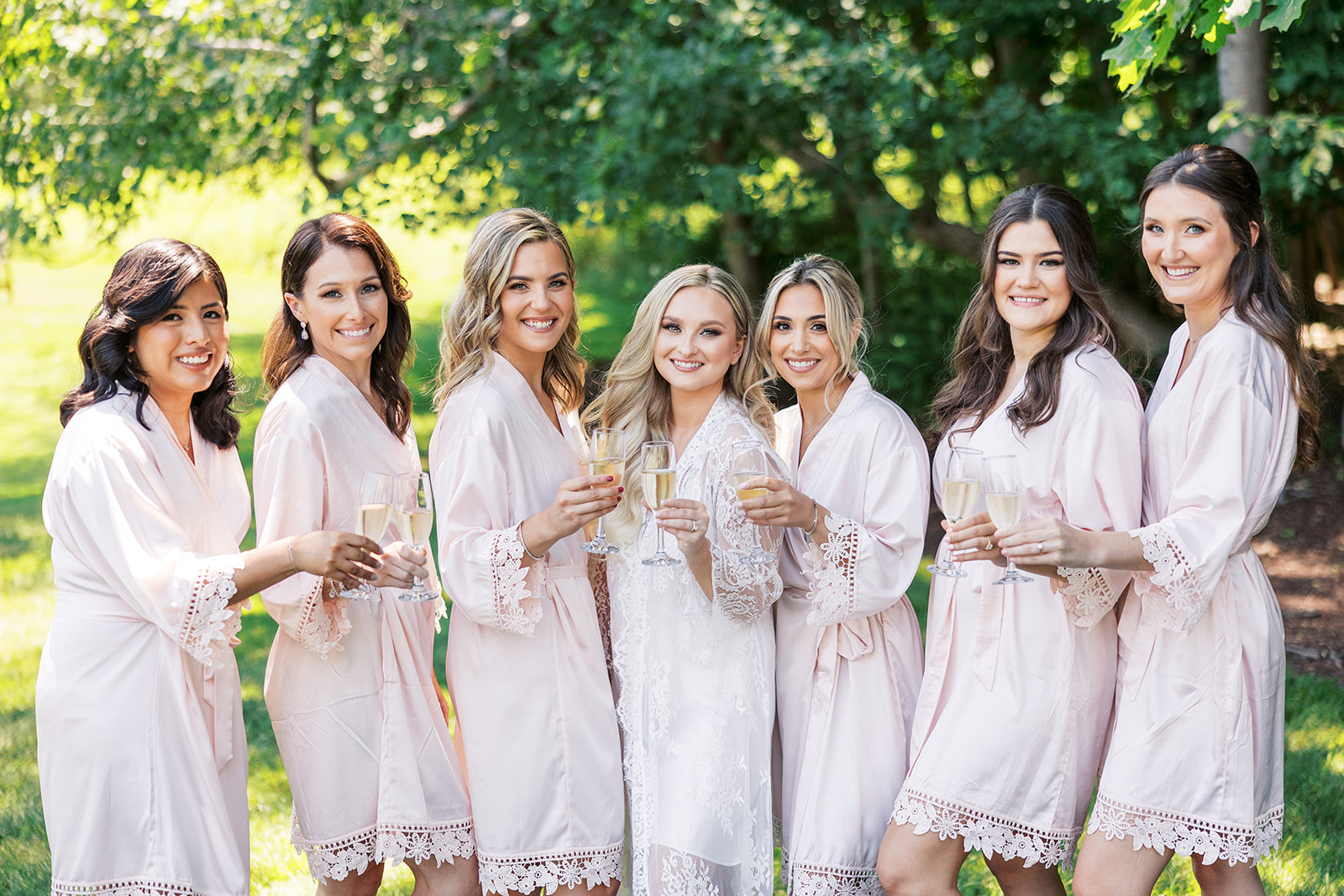 A bride in white pajamas cheers champagne with her bridesmaids in pink pajamas
