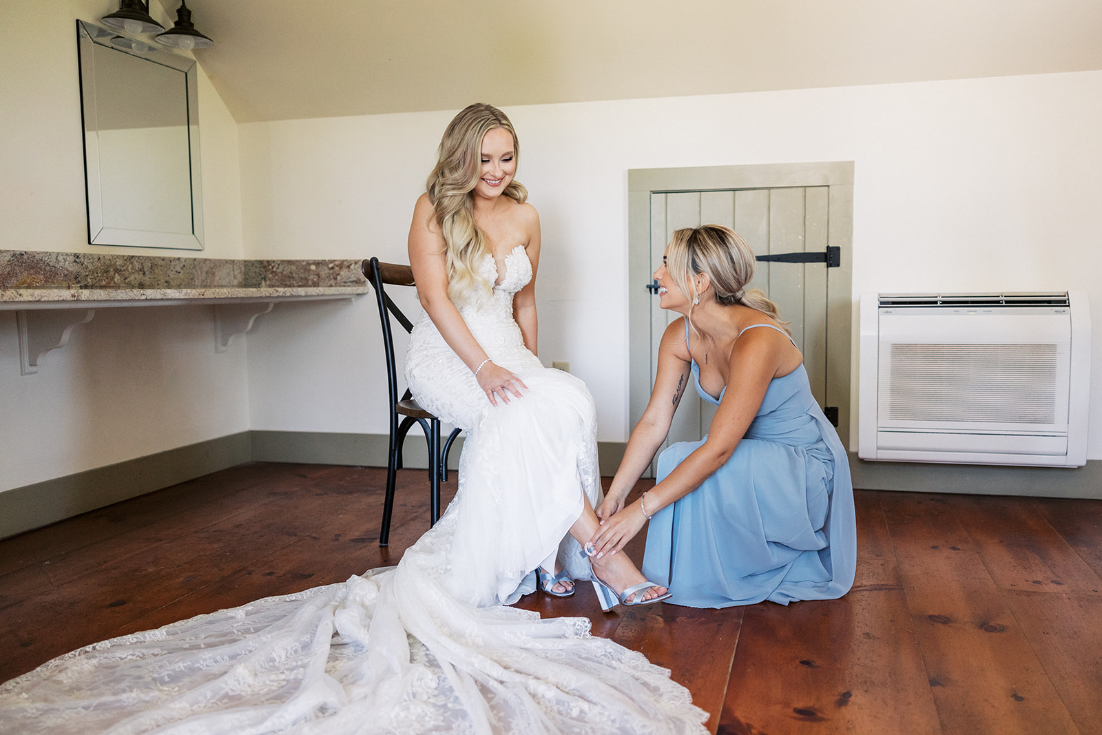 A bridesmaid in a blue dress helps the bride put on her shoes while sitting in a chair