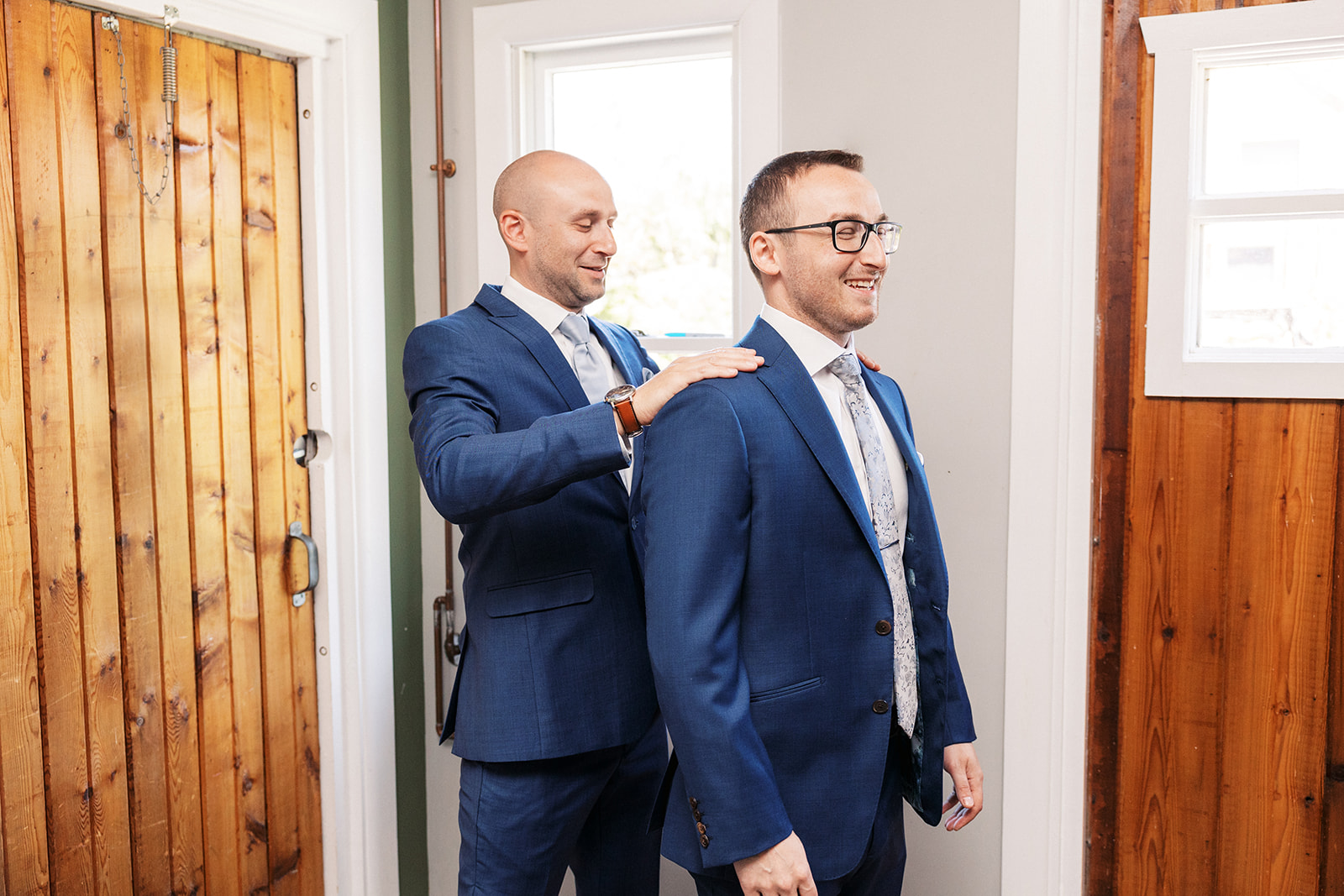 A groomsmen dusts off the shoulders of the groom in blue suits while getting ready for his wedding