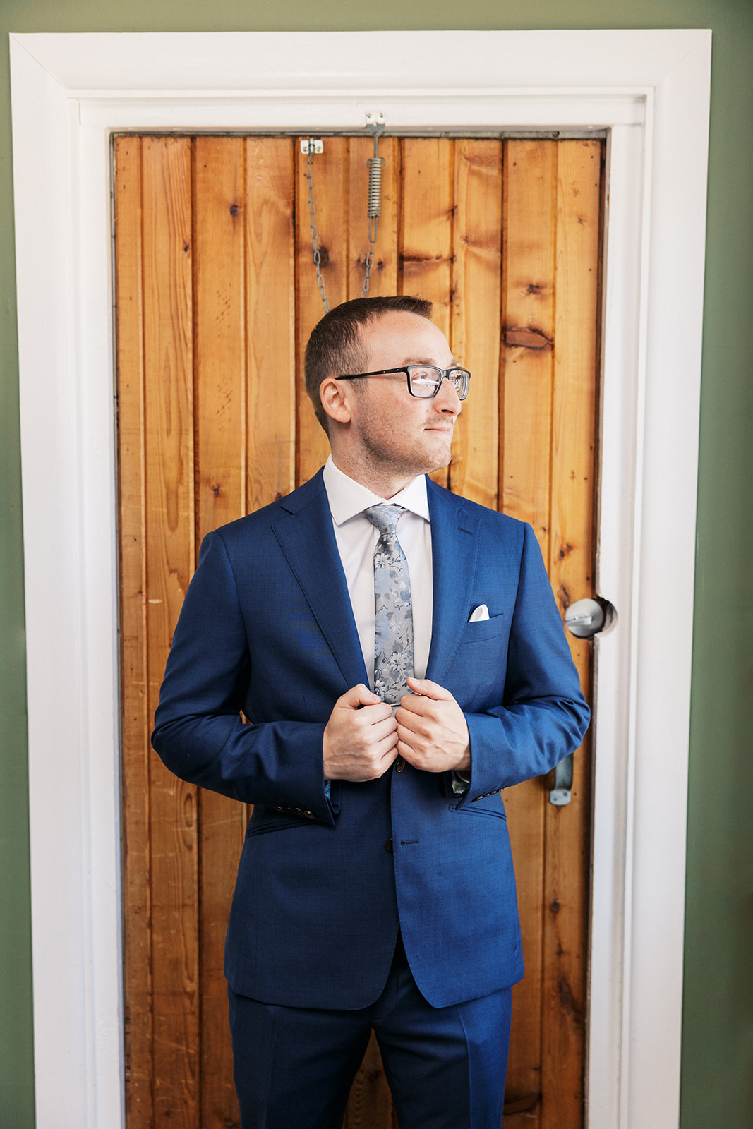 A groom in a blue suit stands in a doorway holding his lapels and looking out a window