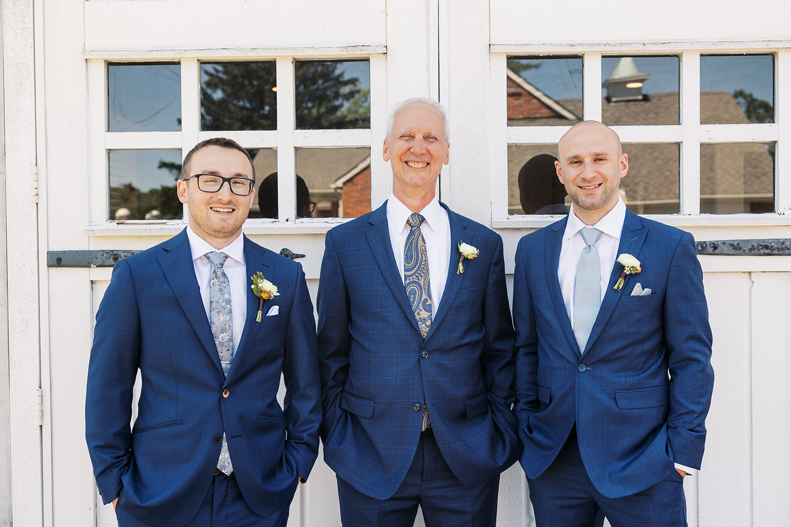 A groom stands in front of white barn doors with his groomsmen in matching blue suits and hands in pockets