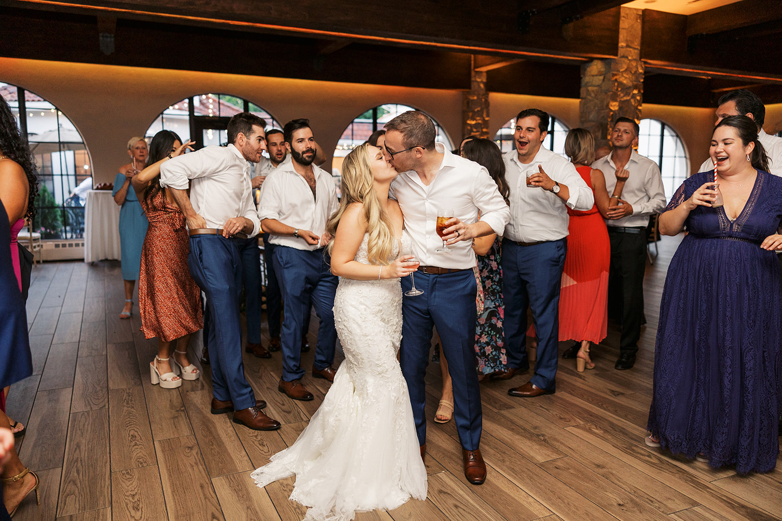 Newlyweds kiss while surrounded by guests holding drinks at their The Reserve At Perona Farms wedding reception