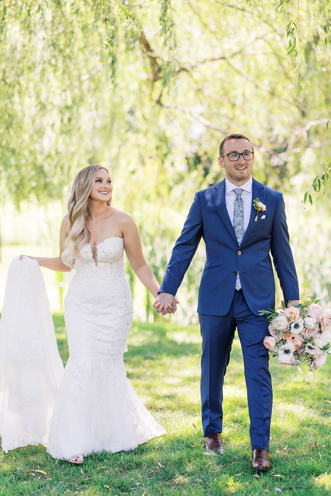 A groom in a blue suit leads his bride through a lawn by the hand while holding her bouquet
