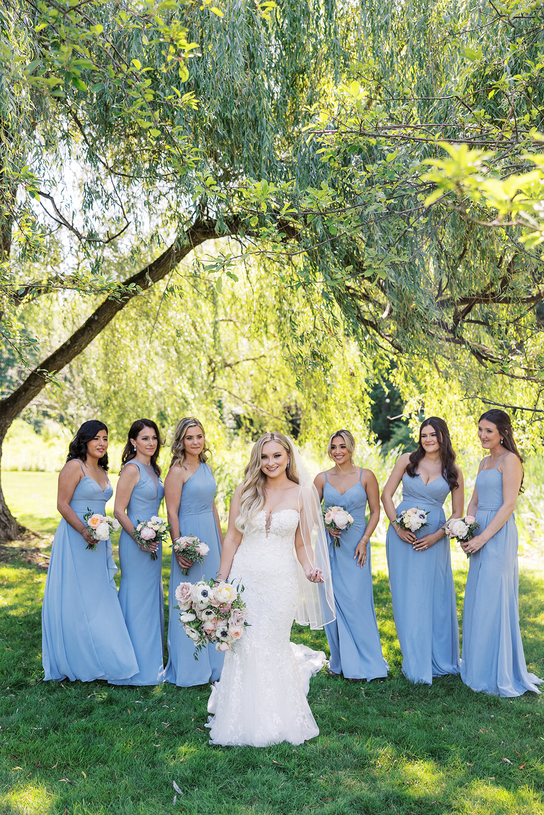 A bride walks under a willow tree in front of her six bridesmaids in baby blue dresses