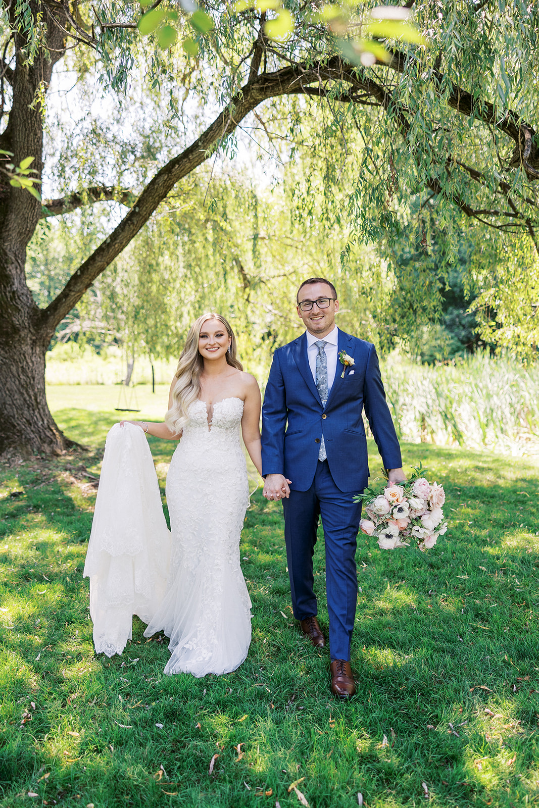 Newlyweds walk under a willow tree holding hands and the dress and bouquet at their The Reserve At Perona Farms wedding