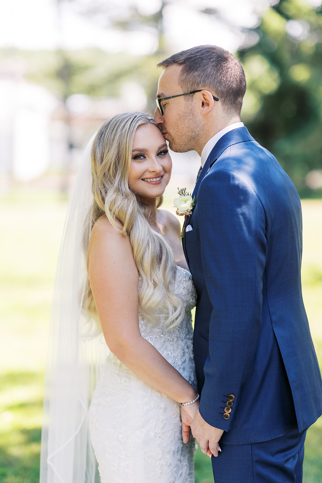 A groom in a blue suit kisses the forehead of his bride while standing in a lawn