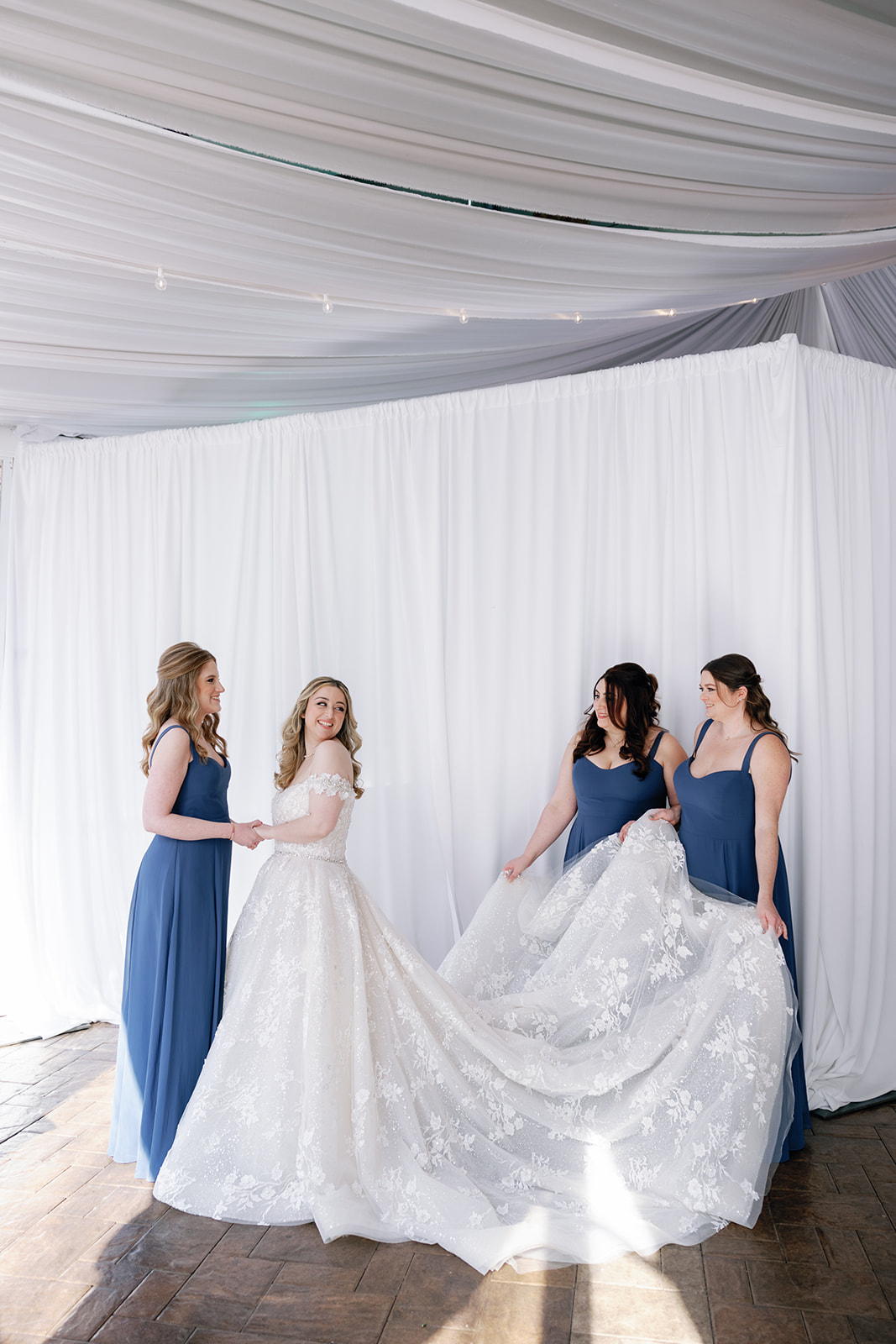 A bride is helped by her bridesmaids in blue dresses with her long lace train