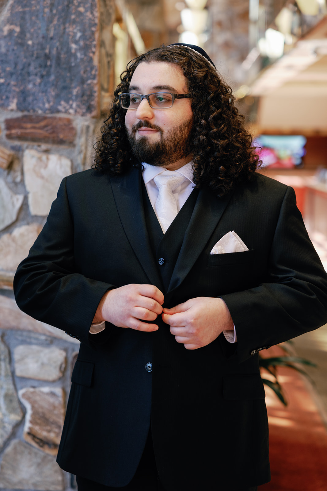 A groom in a black suit buttons his jacket while standing inside