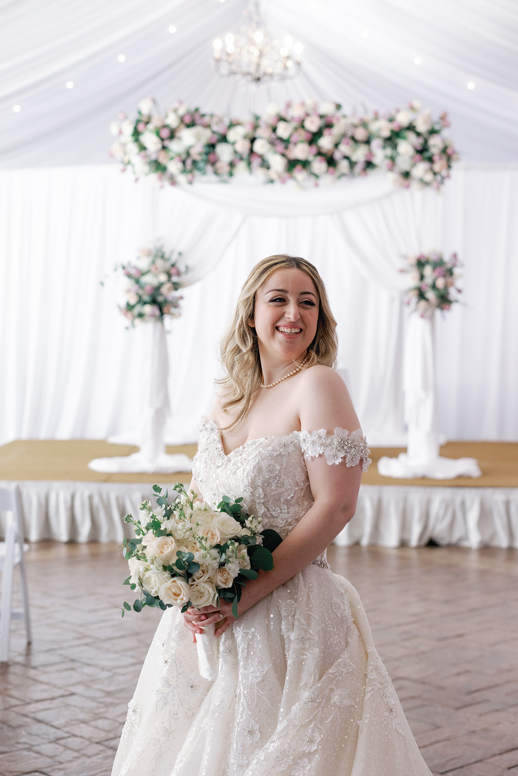 A bride in a lace dress smiles over her shoulder and holding her bouquet in a ceremony room