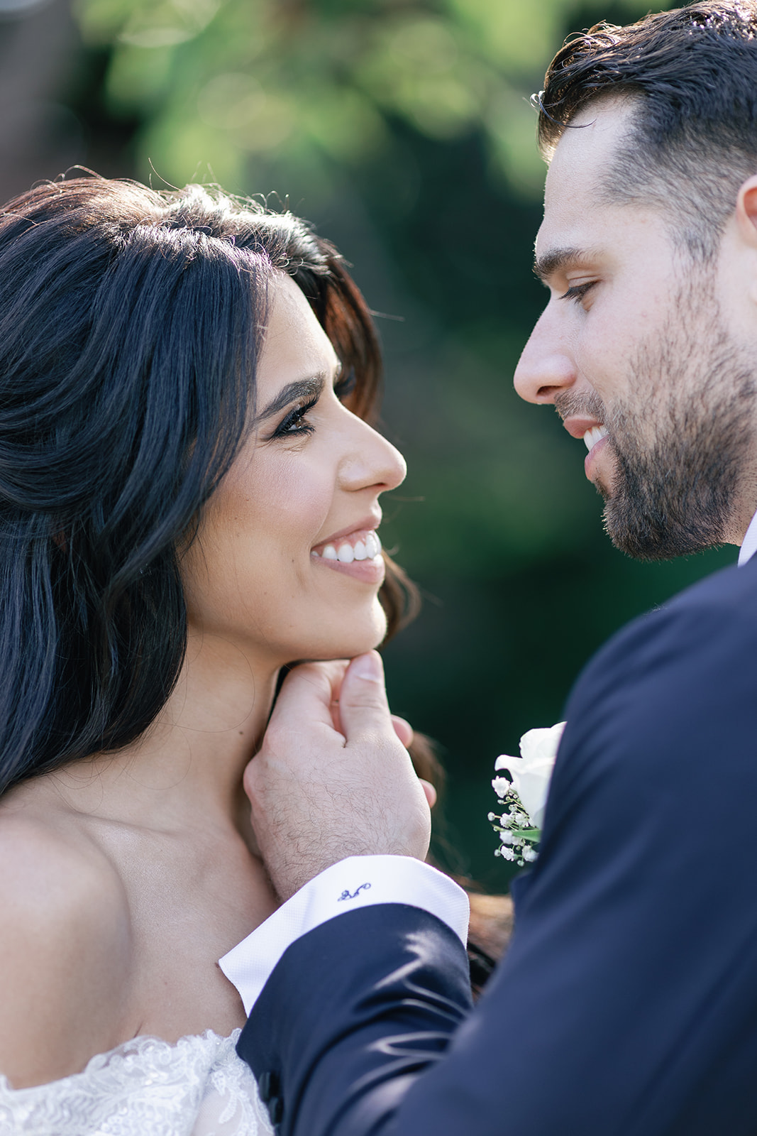 A groom in a black suit touches the chin of his smiling bride while standing in a garden at their Hamilton Manor Wedding