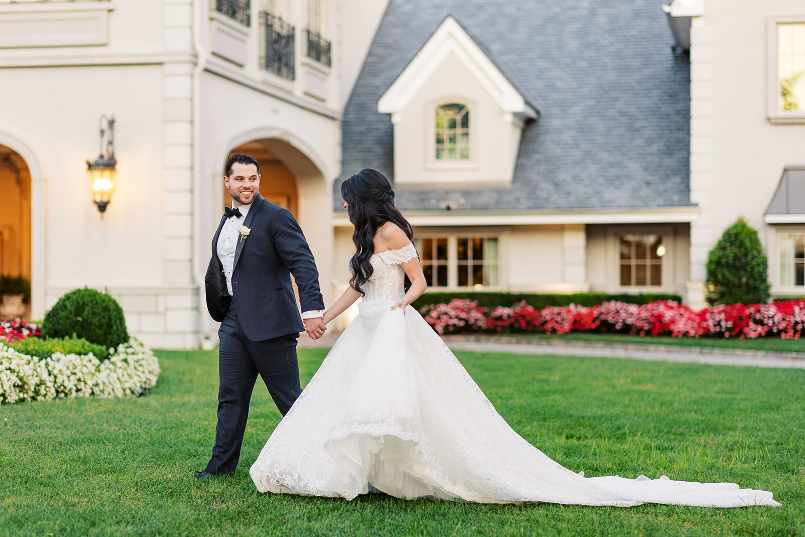 A groom in a black suit leads his bride through a manicured lawn at one of the Castle Wedding Venues in NJ