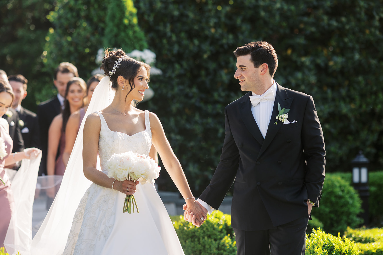 Newlyweds hold hands while smiling at each other and walking through one of the New Jersey Garden Wedding Venues with their wedding party behind them