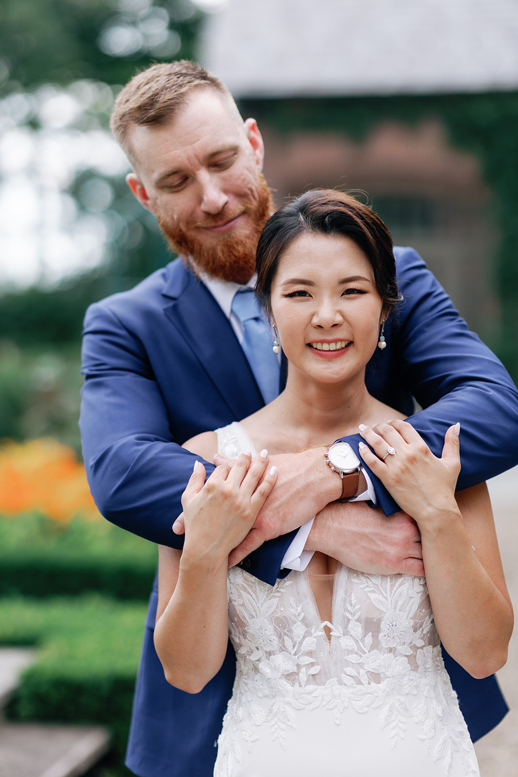 A groom in a blue suit hugs his bride from behind while standing in a garden