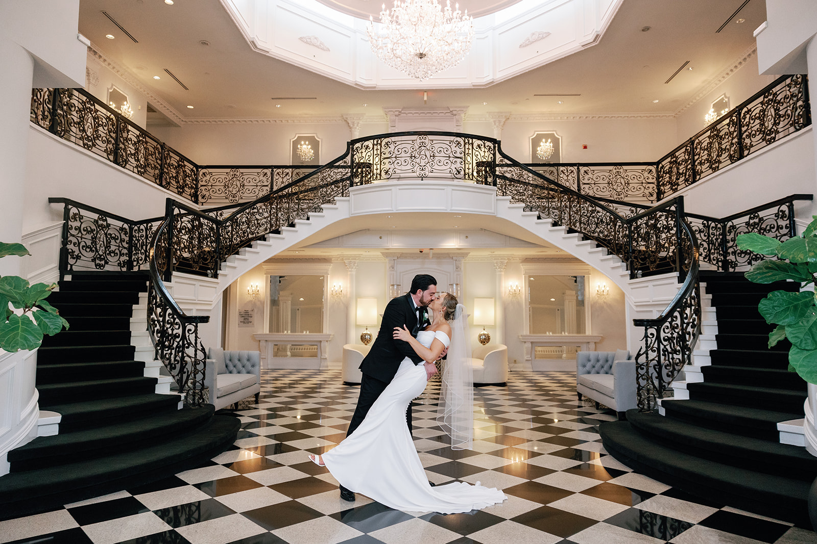 A groom in a black suit dips and kisses his bride under the grand staircase at one of the New Jersey Mansion Wedding Venues