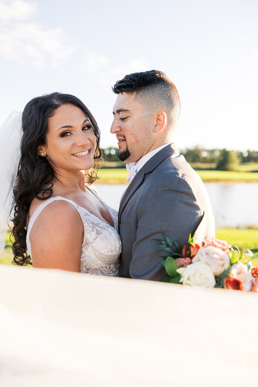 A bride smiles big while being hugged by her new husband on a lakeside lawn at New Jersey Waterfront Wedding Venues