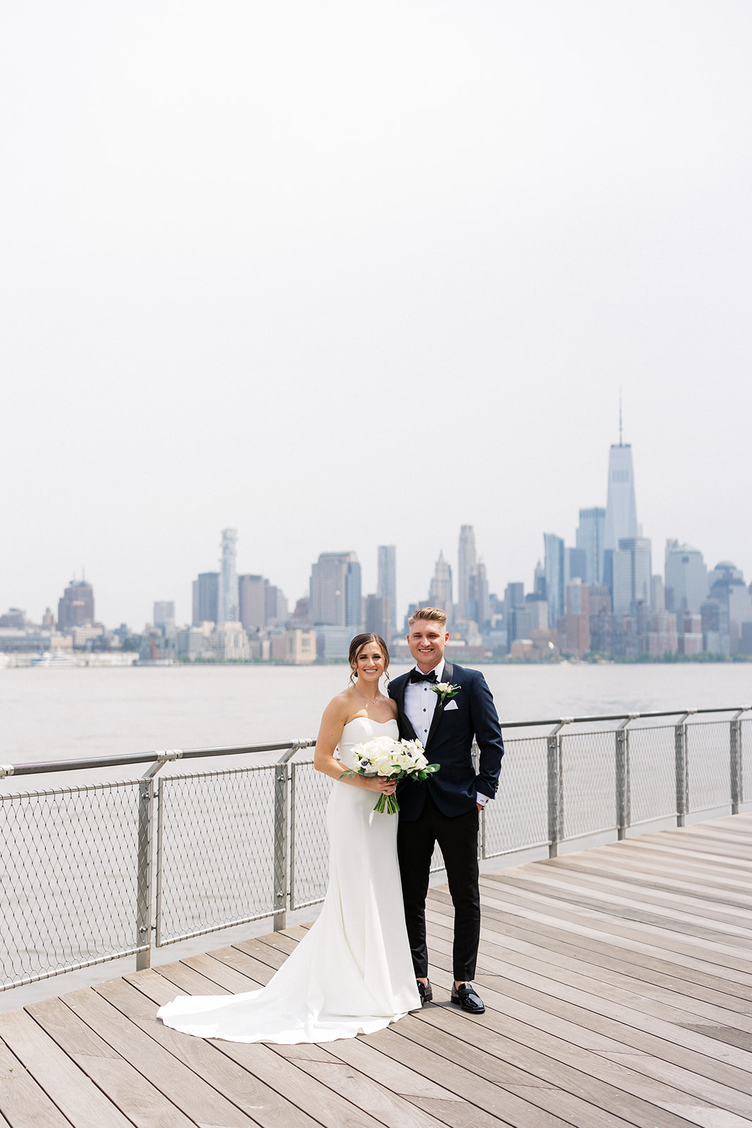 Newlyweds stand on a lakefront park boardwalk with at New Jersey Waterfront Wedding Venues with manhattan skyline across the river