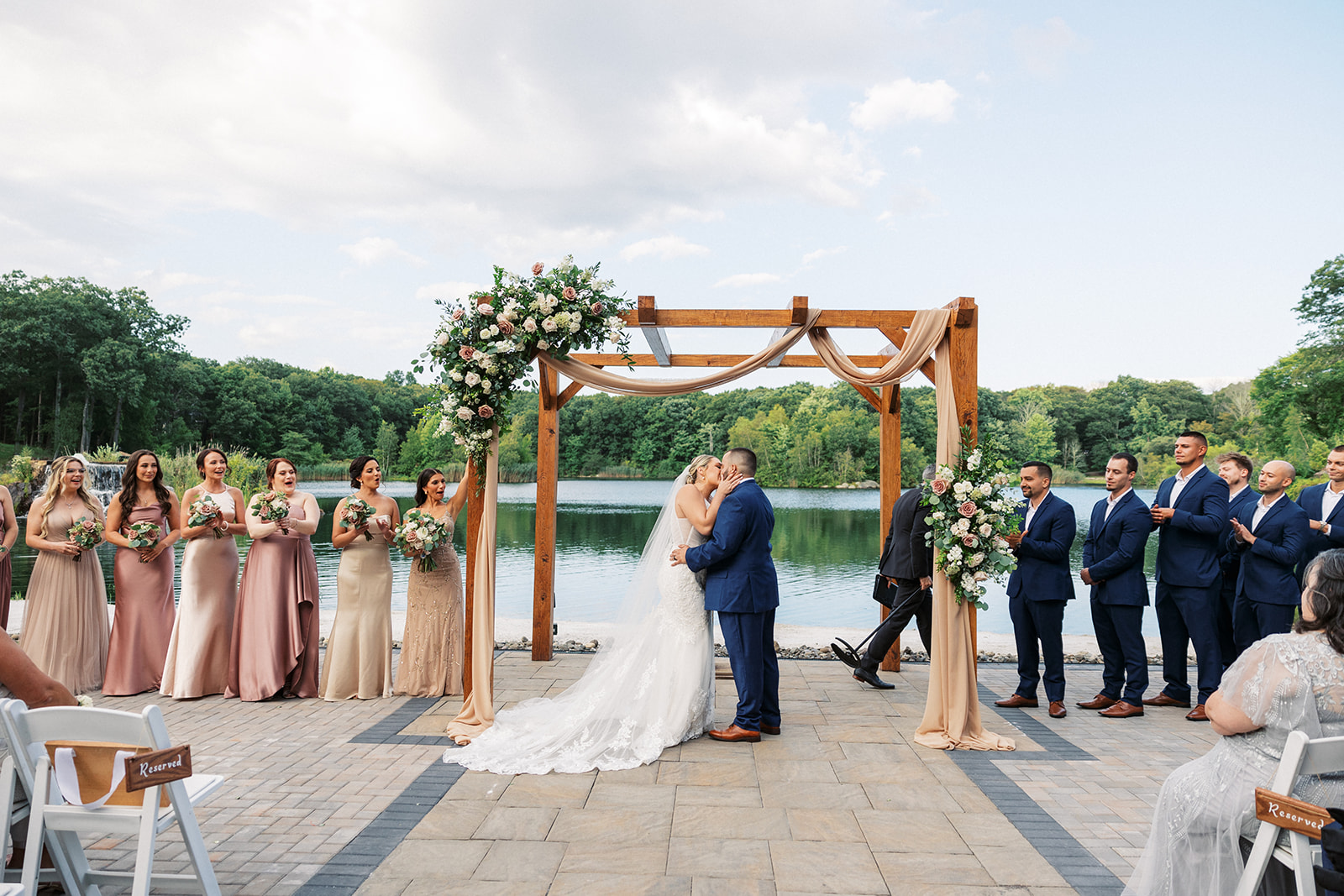Newlyweds kiss under a large wooden arbor surrounded by their wedding party at a lakefront venue
