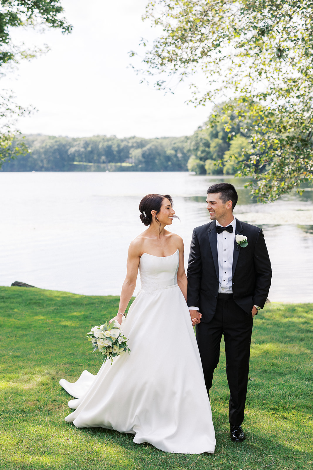 Newlyweds share a big smile while walking hand in hand on a lakeside lawn