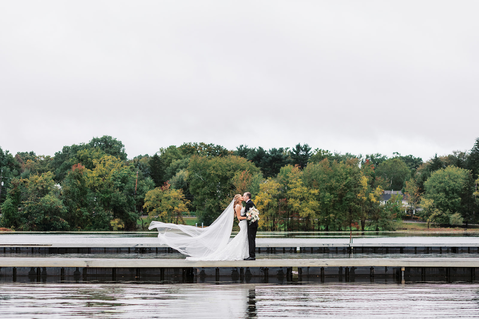 Newlyweds kiss as the veil blows in the wind on the center of a long floating dock in a lake at New Jersey Waterfront Wedding Venues
