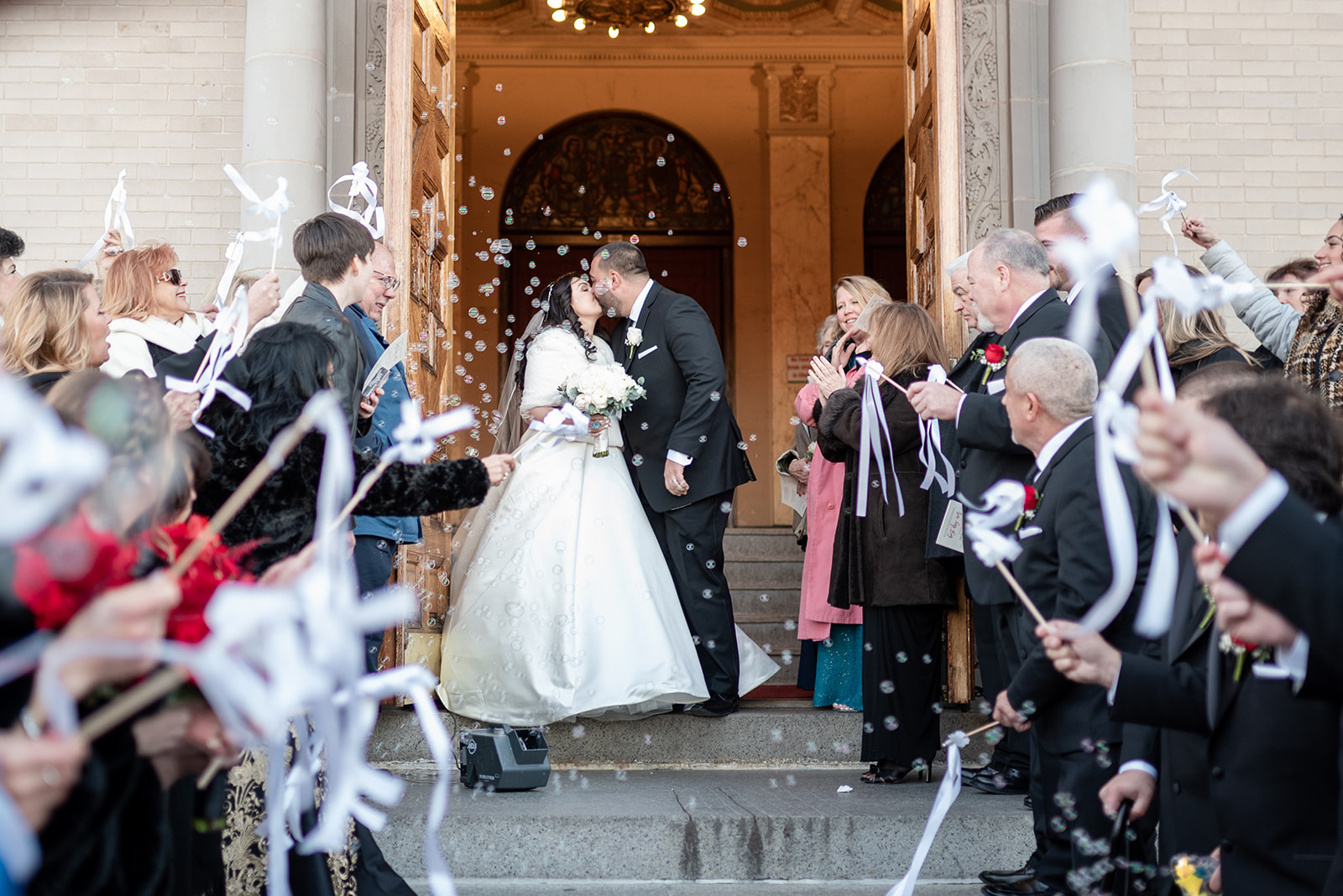 Newlyweds kiss at the exit of their church under a bubble machine and the cheers of their guests on the stairs