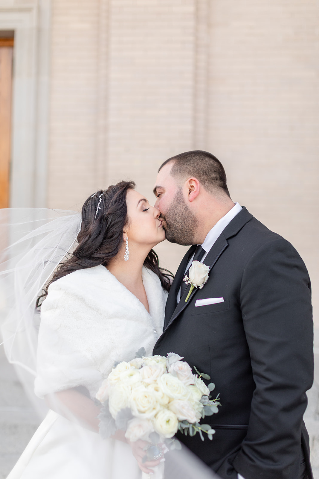 Newlyweds kiss on a windy day on the sidewalk with white roses