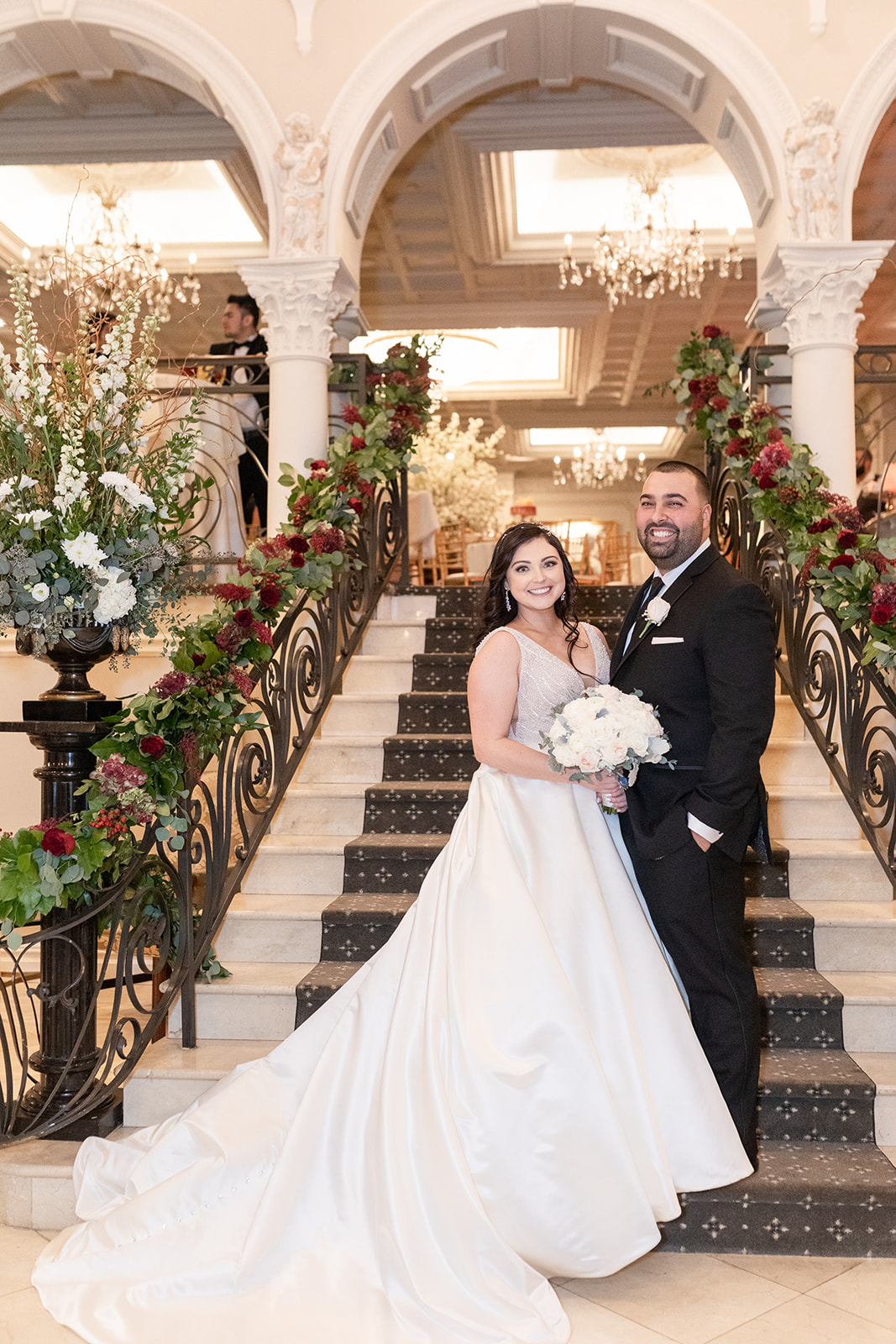 Newlyweds stand on the ornate and red rose covered grand stairs at Nanina’s In the Park wedding venue