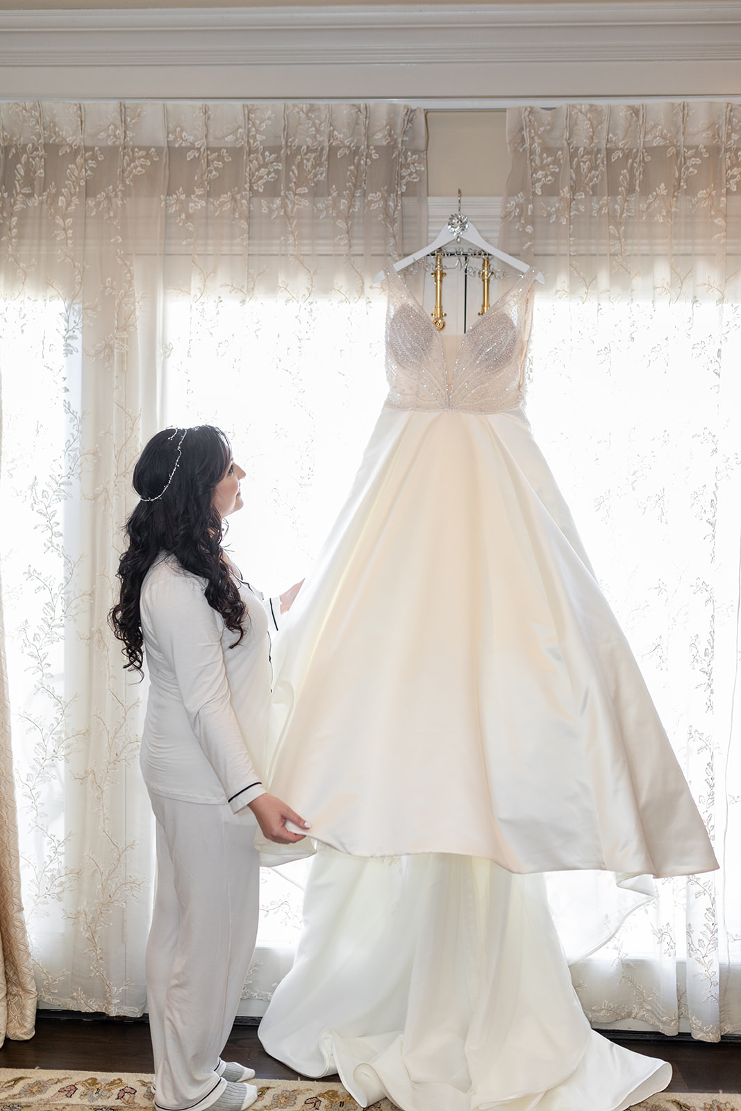 A bride admires her hanging silk dress in a window while wearing white pajamas