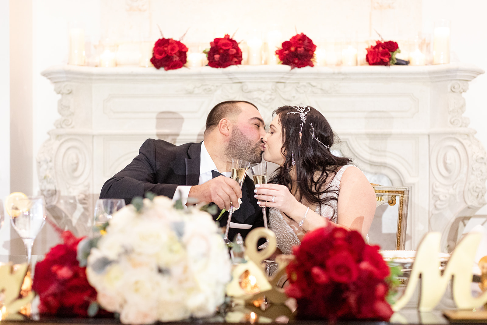 Newlyweds kiss at their rose covered head table in front of the Nanina’s In the Park ornate fireplace