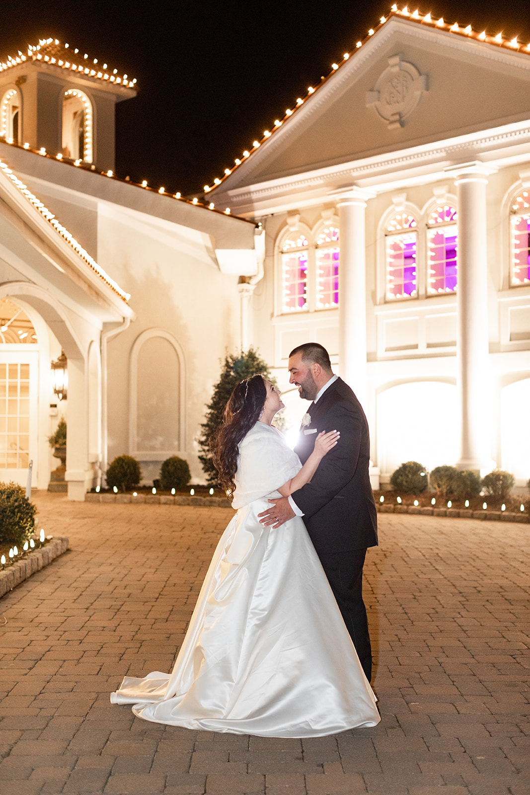 Newlyweds stand smiling at each other in the driveway outside the lit up Nanina’s In the Park wedding venue