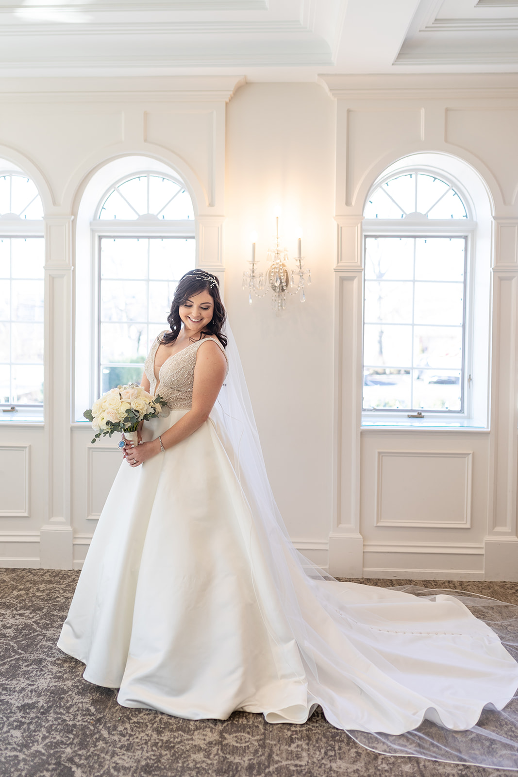 A bride smiles big while standing in her dress and long veil and holding a white bouquet