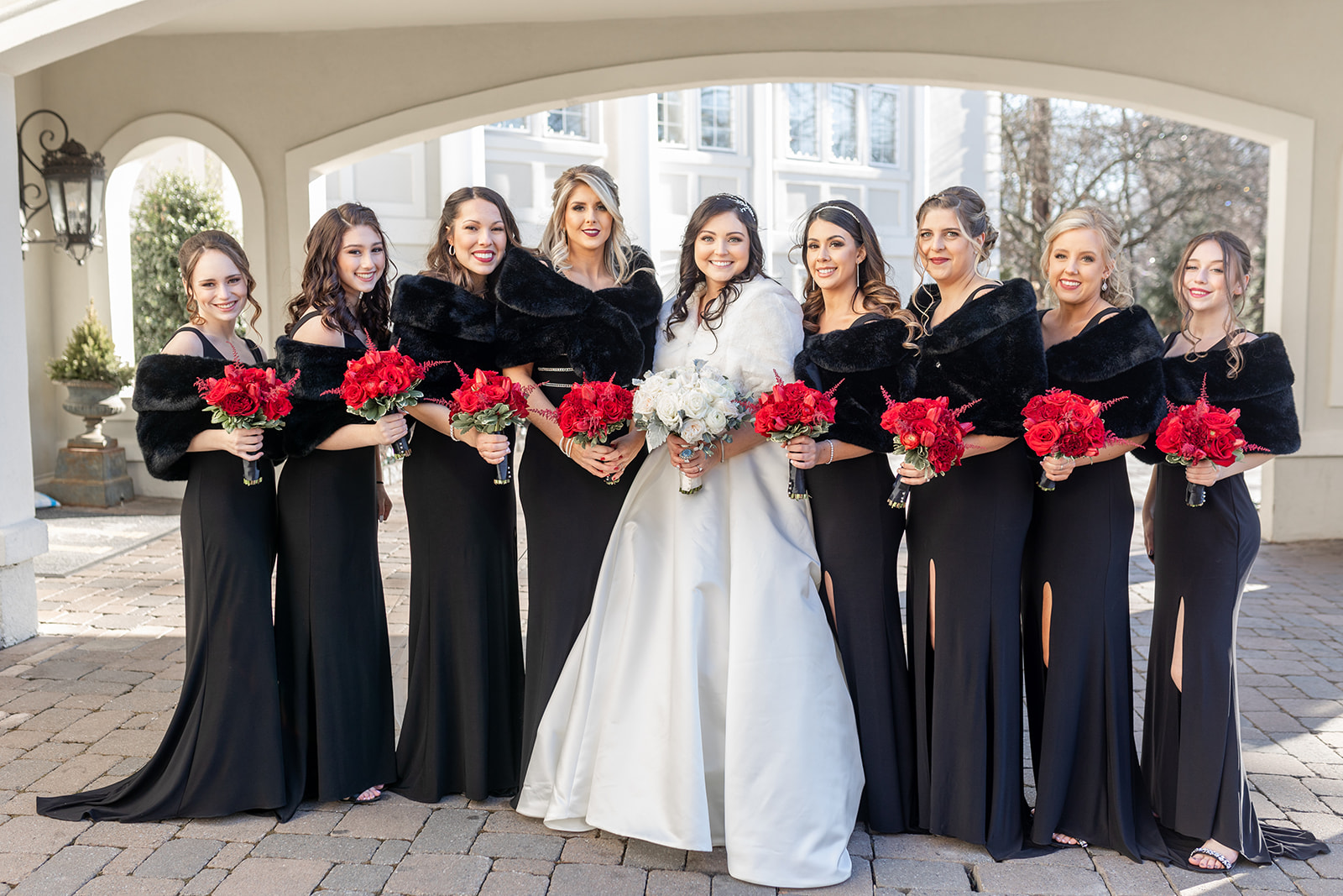 A bride stands in a driveway with her bridesmaids in black dresses holding red and white roses at her Nanina’s In the Park wedding