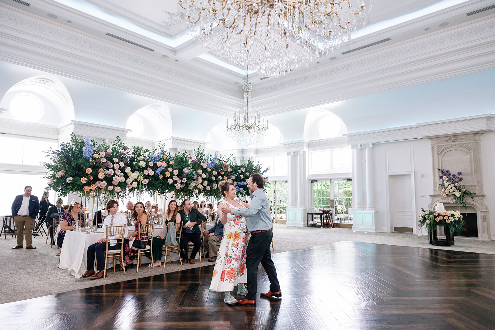 Newlyweds dance for the first time under large crystal chandelier with guests looking on at their Park Chateau Wedding