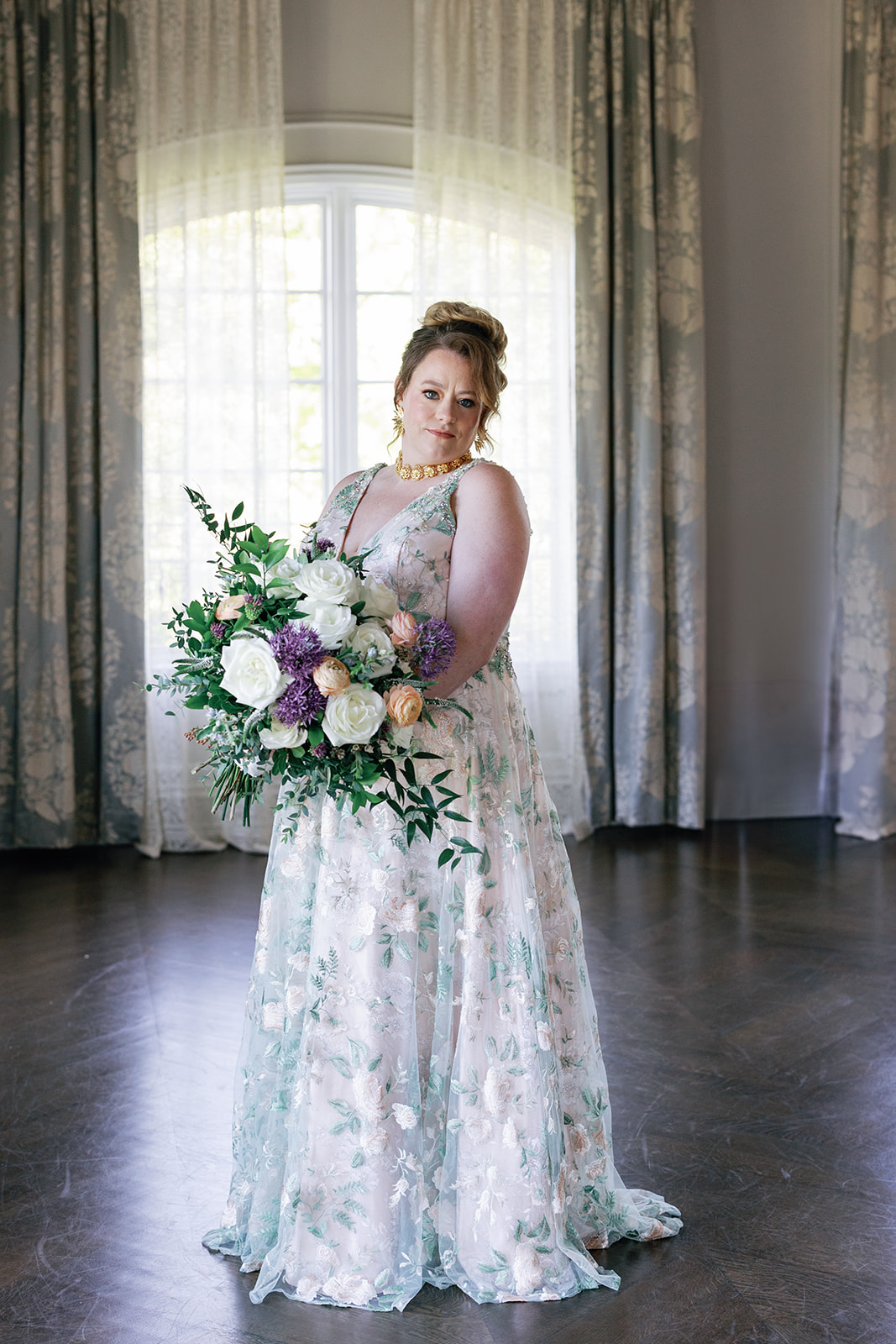 A bride stands in her white and green embroidered dress holding a white and purple bouquet in front of a tall window