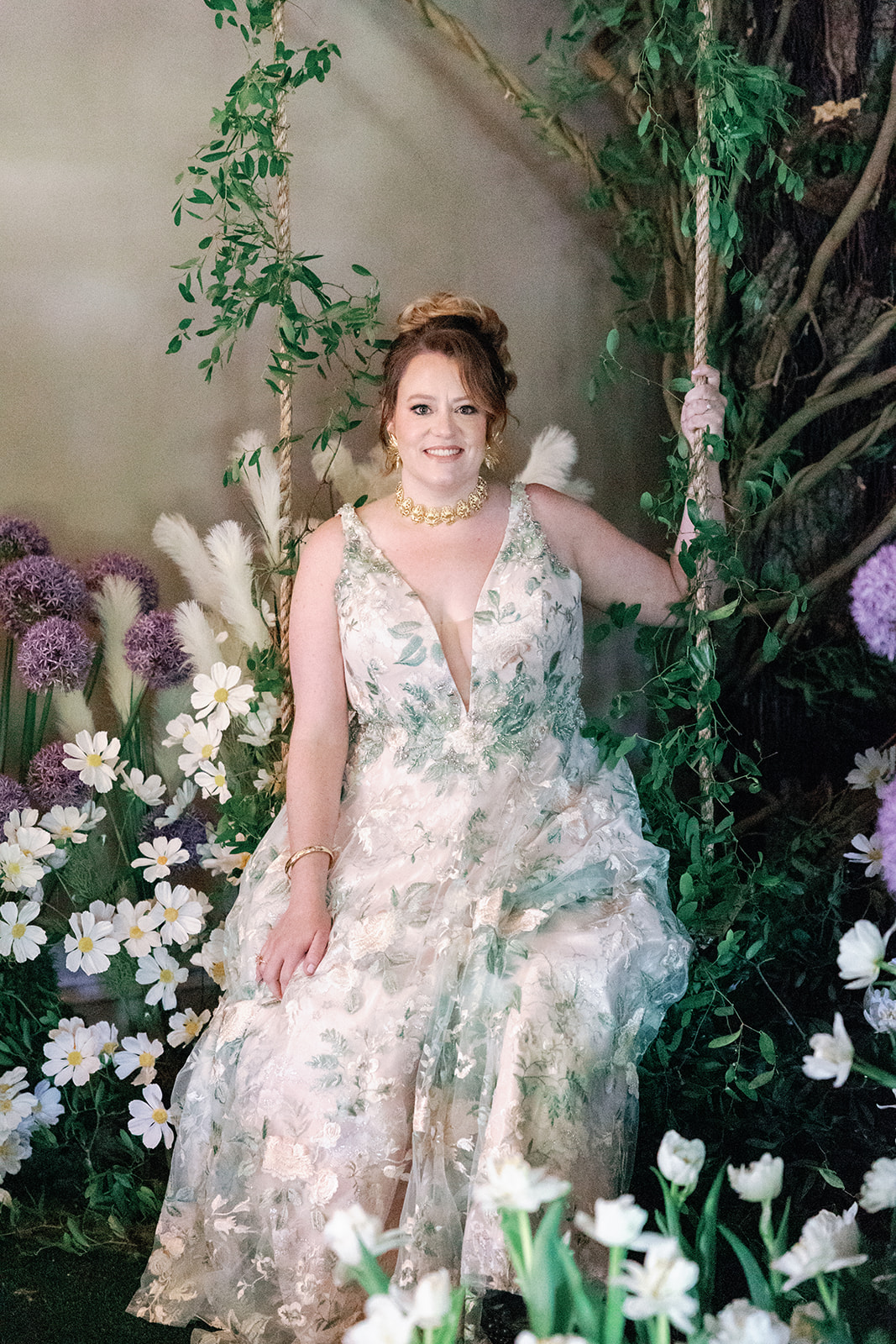 A bride in a green and white dress sits on a swing among many florals at her wedding