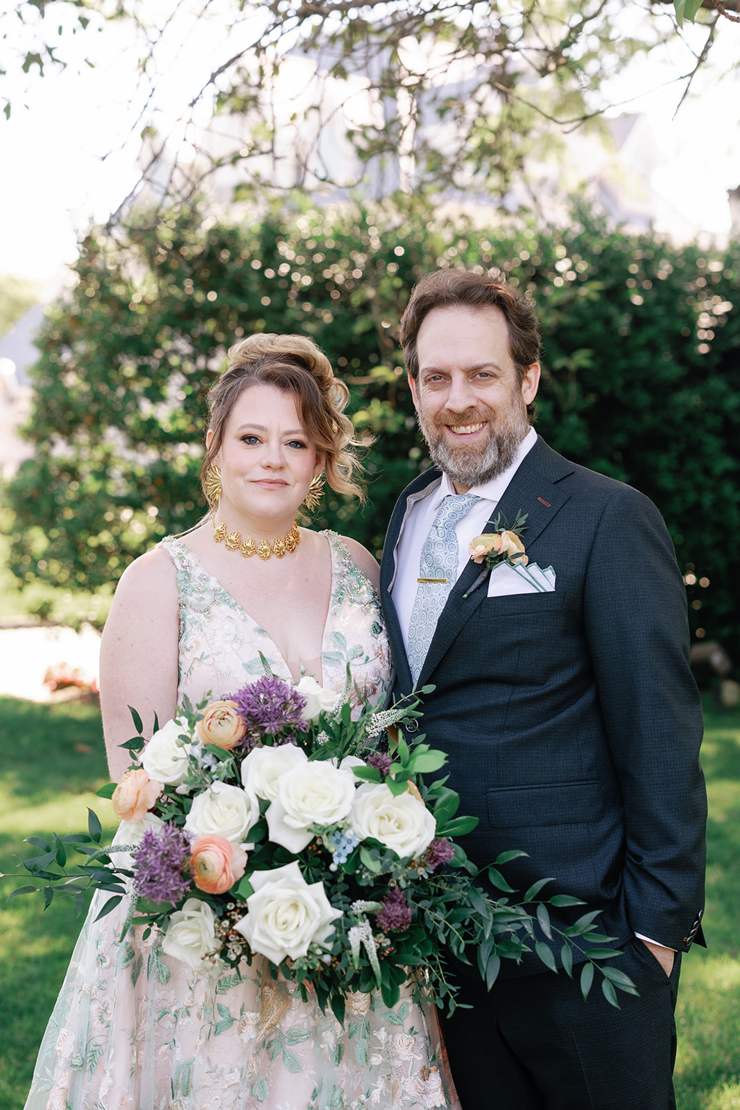 Newlyweds stand under a tree in the gardens holding each other and the large bouquet