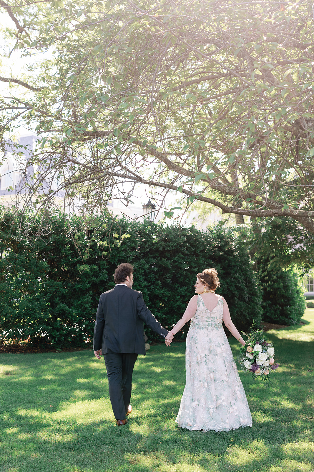 A newlywed couple walks through the gardens holding hands under a tree