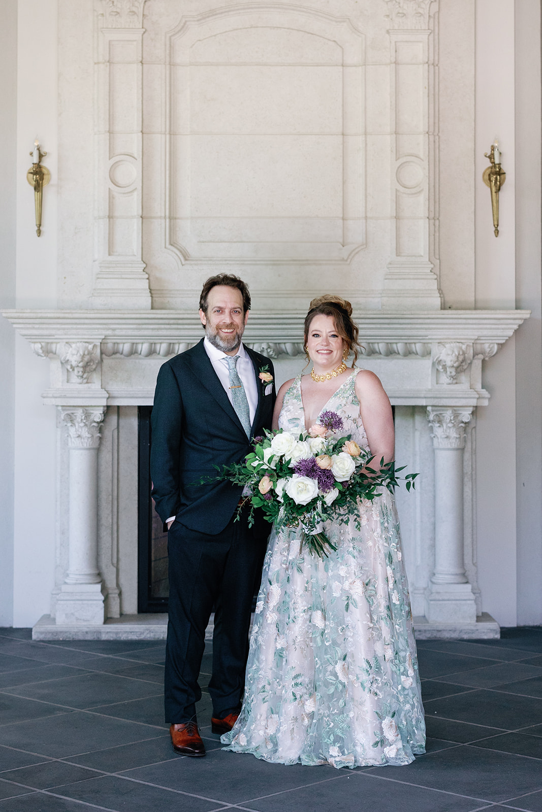 Newlyweds stand in front of an ornate fireplace with a bouquet and smiling
