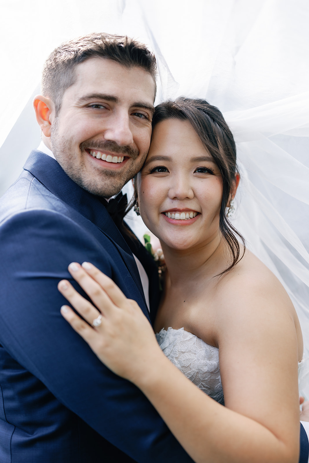 Newlyweds smile while hugging under the large veil during their day at The Grove Wedding Venue